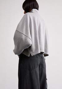Sudadera gris corta con cuello alto y hombros caídos, combinada con pantalones negros de cintura alta que cuentan con bolsillos traseros y una textura sólida.