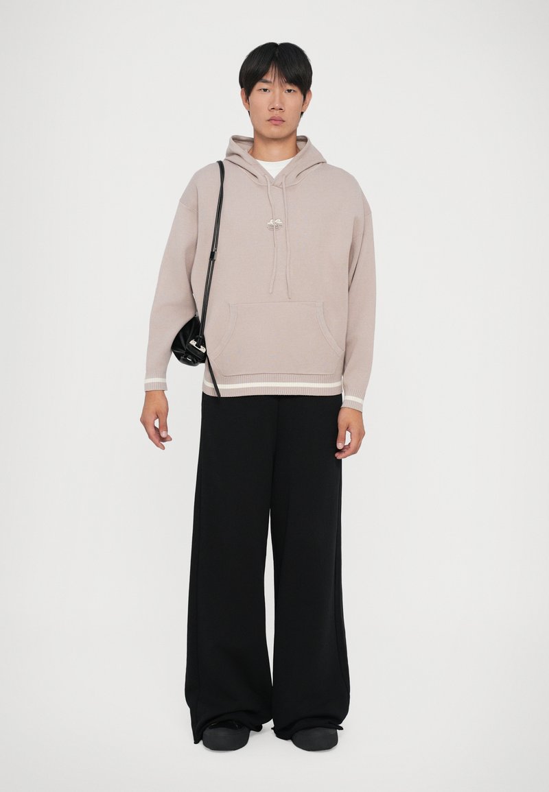 Junger Mann, der einen beigen Hoodie, weite schwarze Hosen, schwarze Schuhe und eine schwarze Umhängetasche auf weißem Hintergrund trägt.