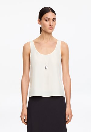 Femme aux cheveux foncés attachés, portant un haut blanc sans manches, une jupe noire et un long collier pendentif argenté, debout sur un fond blanc.