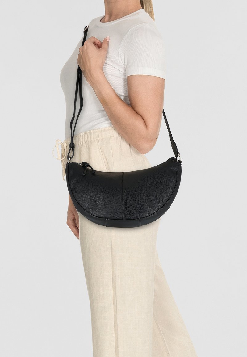 Bolso de mano de cuero negro con una forma curva y en media luna. Presenta una correa trenzada y un acabado texturizado. Combinado con pantalones de color claro.