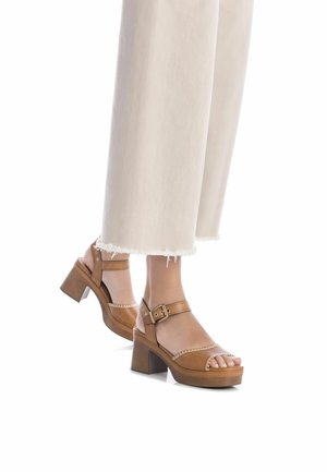 Sandalias de plataforma de cuero marrón con una correa ajustable en el tobillo, punta abierta y un tacón de madera texturizada. Llevadas con pantalones claros deshilachados.