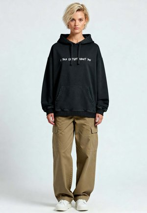 Felpa nera oversize con testo bianco "TI HO PARLATO DI CH TGBT," abbinata a pantaloni cargo beige e sneakers bianchi.