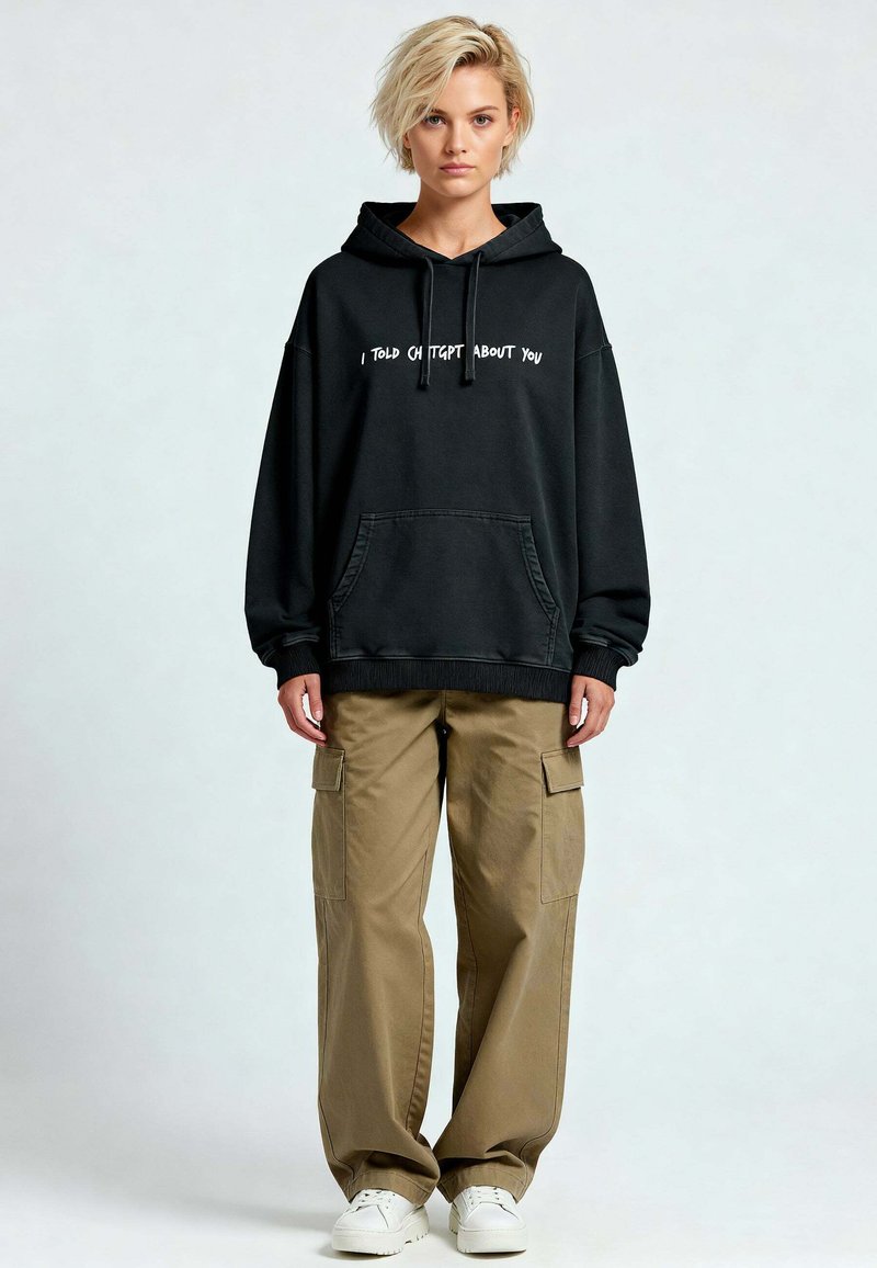 Felpa nera oversize con testo bianco "TI HO PARLATO DI CH TGBT," abbinata a pantaloni cargo beige e sneakers bianchi.