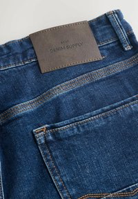 Vaqueros de mezclilla azul oscuro con un parche de cuero marrón en la cintura con texto en relieve, costuras naranjas contrastantes y un bolsillo trasero.