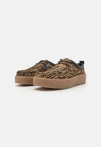 Slip-on sko med et zebra mønster i svart og gyllenbrun på en strukturert overflate, med en kraftig beige såle og svarte detaljer.