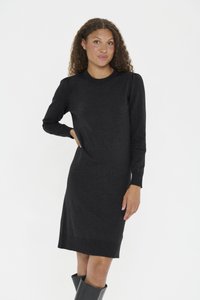 Göndör hajú nő hosszú ujjú fekete kötött ruhában és fekete térdig érő csizmában, egyszerű világos háttér előtt állva.