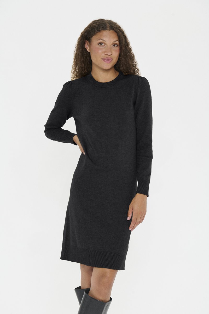 Göndör hajú nő hosszú ujjú fekete kötött ruhában és fekete térdig érő csizmában, egyszerű világos háttér előtt állva.