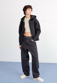 Fekete cipzáras kapucnis pulóver egy bézs cropped toppal, laza fekete farmernadrággal párosítva. Világos színű sportcipők egészítik ki az outfitet. Semleges háttér.