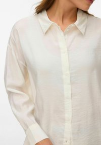 Femme portant une chemise ample à manches longues et boutons, de couleur blanc cassé, avec un col, montrée du torse au cou.