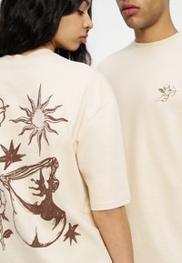 Δύο άτομα που φορούν κρεμ μπλουζάκια· μια μπλούζα έχει έναν καφέ ήλιο, αστέρια και μια κλασική προτομή στην πλάτη, ενώ η άλλη έχει έναν μικρό αγγελούδα με τόξο στο μπροστινό μέρος.