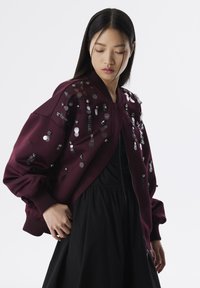 Chaqueta bomber burdeos con lentejuelas redondas dispersas y detalles en negro, con puños acanalados y cierre de cremallera. Usada sobre un vestido negro.