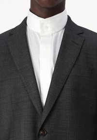 Kulkraft grå blazer med et struktureret stof, enkeltknaplukning og en hvid skjorte med høj krave under. Ingen synlige mønstre eller detaljer.