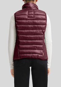 Person, der eine ärmellose kastanienbraune Steppweste über einem weißen langärmeligen, gerippten Shirt und schwarzen Hosen trägt, von hinten gezeigt.