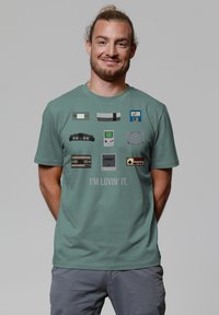 Groene katoenen T-shirt met retro gameconsole graphics; blokkige vormen, lichte kleuren, en de tekst "I'M LOVIN' IT" in het midden.