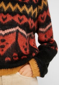 Gros plan d'une main dans la poche d'un pantalon beige portant un pull en tissu pelucheux avec des motifs géométriques et de cœurs rouges, noirs, oranges et jaunes.
