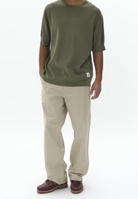 Olivově zelené tričko s krátkým rukávem, béžové volné kalhoty a hnědé kožené lodní boty s tkaničkami. Neformální styl, minimální značení na tričku.