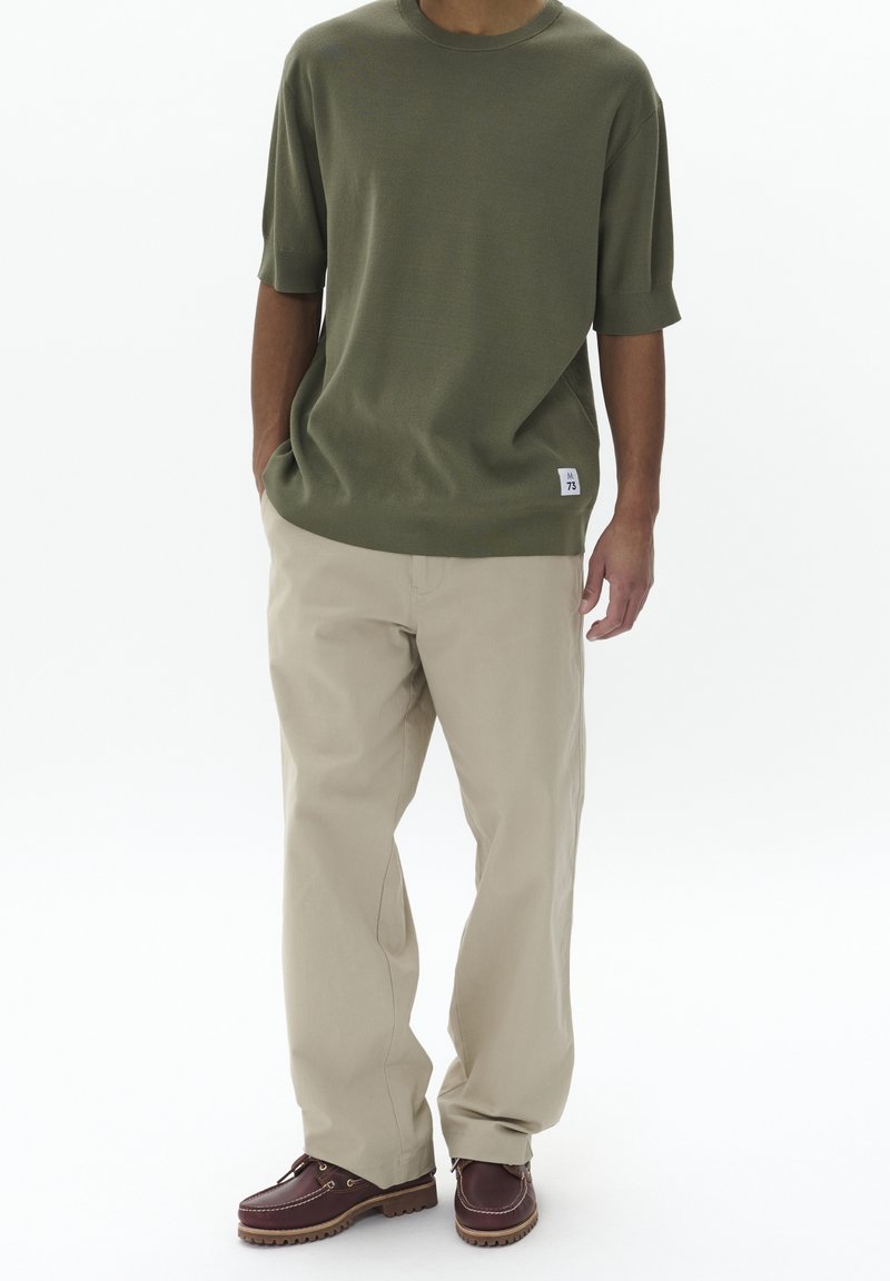 Olivově zelené tričko s krátkým rukávem, béžové volné kalhoty a hnědé kožené lodní boty s tkaničkami. Neformální styl, minimální značení na tričku.