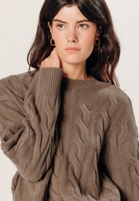 Brauner Kabelstrickpullover mit Rippkragen und oversized Passform. Der Stoff wirkt weich mit einem strukturierten Muster und entspannten Ärmeln.
