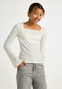 Langærmet top i hvid ribbet stof med firkantet udskæring og udvidede ærmer, parret med grå denim jeans. Teksturfinish synlig.