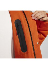 Orange Jacke aus strukturiertem Stoff, mit einer schwarzen Reißverschluss-Tasche auf der Brust und einem sichtbaren Reißverschluss auf der Vorderseite.