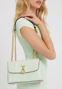 Mátová zelená kabelka s krokodýlí texturou, zlatými detaily, černými akcenty a řetízkem, kterou drží osoba v mátovém topu s ribkováním.