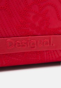 Rote Ledertasche mit strukturierten Stickmustern und einem auffälligen, geprägten "Desigual"-Label an einem glatten Tragegurt.