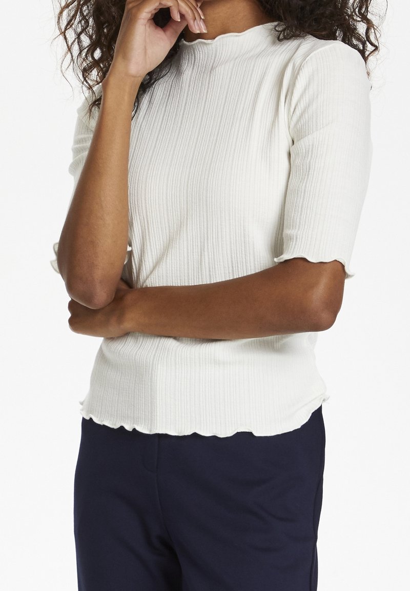 Top bianco a maniche corte con righe e scollatura e orlo a scallop, caratterizzato da un design aderente. Indossato con pantaloni blu navy.