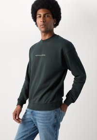 Sweatshirt vert foncé à col rond et poignets côtelés ; présente un logo imprimé blanc "SCOTCH & SODA" sur la poitrine. Porté avec un jean bleu clair.