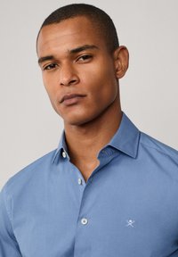 Camisa de botones en azul claro, hecha de algodón. Presenta un cuello, botones blancos y un sutil logo bordado en el pecho.