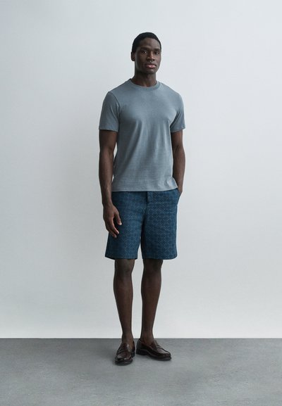 Νεαρός άνδρας στέκεται μπροστά σε απλό φόντο, φοράει ανοιχτό μπλε μπλουζάκι, ναυτικά σορτς με σχέδιο και καστανά σκουροκαφέ loafers.