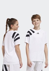 T-shirt de sport blanc avec des rayures noires sur les épaules, à manches courtes et avec un logo. Tissu léger et coupe décontractée conçue pour les vêtements de sport.