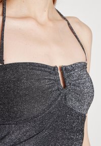 Iskrzący, ciemnoszary top na halter z małym detalem w kształcie klucza z przodu, noszony przez osobę, eksponujący ramiona i górną część klatki piersiowej.