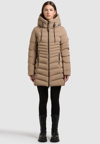 Beżowa pikowana kurtka puchowa z kapturem, pionowym pikowaniem, zamkiem z przodu i czarnymi detalami. Noszona z czarnymi legginsami i butami.