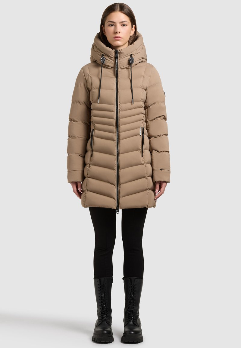 Beżowa pikowana kurtka puchowa z kapturem, pionowym pikowaniem, zamkiem z przodu i czarnymi detalami. Noszona z czarnymi legginsami i butami.