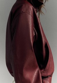 Bordowa skórzana kurtka z wysokim kołnierzem, opadłymi ramionami i zamkiem błyskawicznym z przodu. Wyjątkowa kieszeń boczna i gładka faktura.