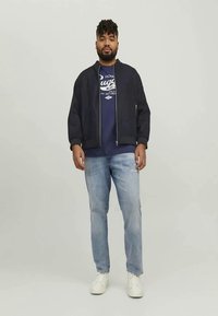 Veste aviateur bleu marine avec fermeture éclair sur le devant, poignets côtelés, portée sur un T-shirt graphique, associée à un jean bleu clair et des baskets blanches.