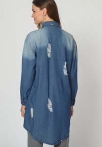 Vestido de camisa de mezclilla en azul medio con efecto desgastado, con parches desgastados y dobladillo curvado, mangas largas con puños y cuello tipo camisa.