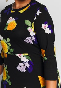 Robe noire ornée de roses jaunes, de fleurs violettes et blanches. Présente un col rond et des manches trois-quarts. Tissu doux et ajusté.