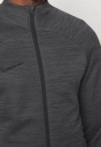 Nike zipová bunda v tmavě šedé barvě, se strukturovaným vzorem, vysokým límcem a černým zipem. Na hrudi se nachází malé logo.