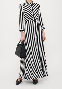 Kobieta w długiej sukience w czarno-białe paski trzyma czarną torebkę, a u stóp ma czarne, siateczkowe buty Mary Jane.