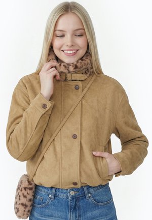 Smilende kvinde med langt blond hår iført en tan farvet jakke med knapper, kunstpels halsedisse, crossbody-taske og blå jeans mod en hvid baggrund.