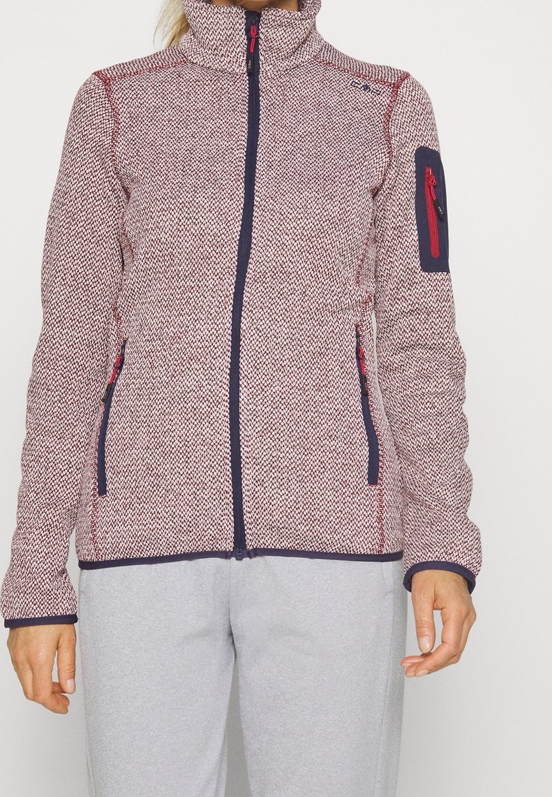 Femme portant une veste zippée texturée rouge et blanche avec bordures bleu marine et poches zippées, associée à un pantalon gris clair.