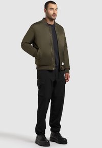 Olivgrüne Bomberjacke mit Reißverschluss, gerippten Bündchen und zwei seitlichen Taschen, kombiniert mit schwarzen Cargo-Hosen und schwarzen Schnürstiefeln.