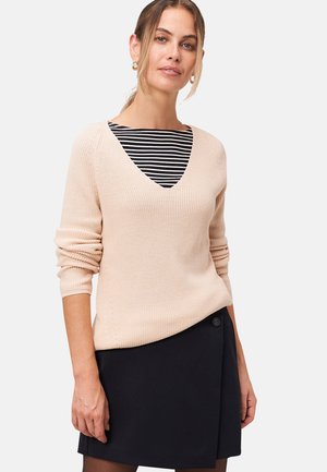 Pull en tricot beige clair avec un décolleté en V, associé à un top à rayures noir et blanc. Jupe noire avec un détail de bouton.