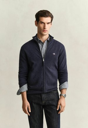 Man wearing a navy zip-up sweater over a gray striped shirt with rolled-up sleeves and dark jeans standing against a plain background.