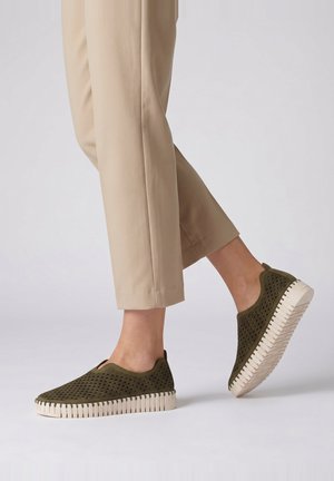 Person wearing beige cropped pants and olive green slip-on shoes with white textured soles, standing on one foot in neutral background.