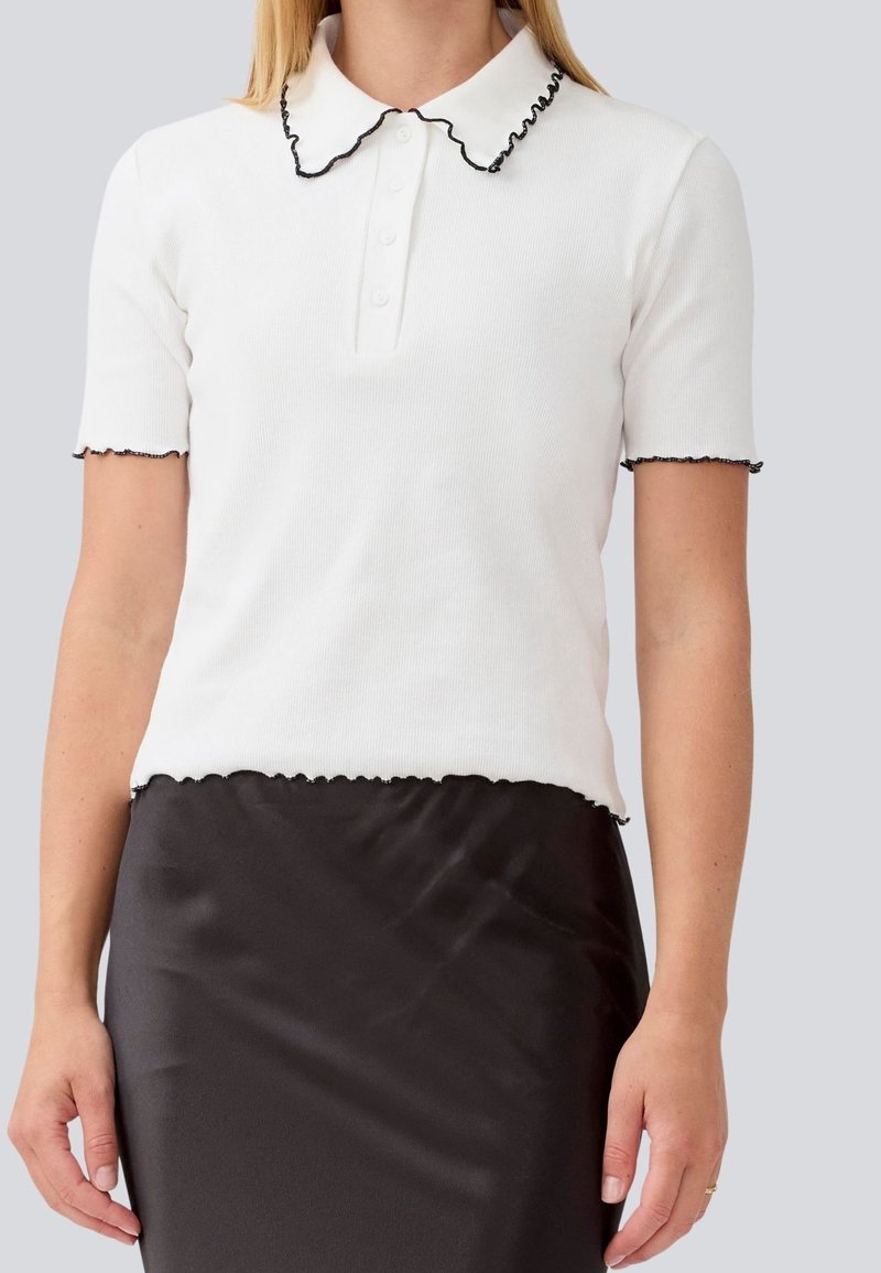 Femme portant un polo blanc à manches courtes avec des bords noirs volantés et une jupe noire sur un fond uni.
