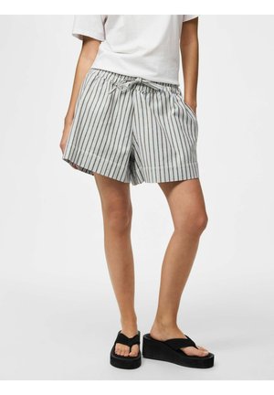 Person som har på seg hvite stripete shorts med snøring og lommer, hvit t-skjorte og svarte platåsandaler står mot en ensfarget bakgrunn.