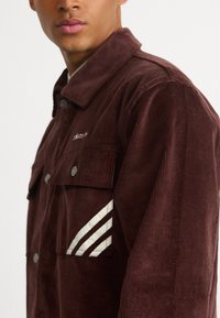 Pruunist korduroožaketist, millel on nööbiga esiosa, kaks rinnataskut ja valge kolme triibuga aktsent varrukal. Tekstureeritud pind.