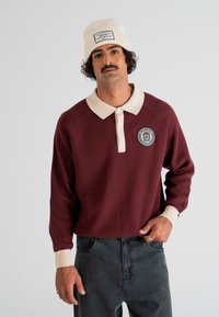 Man with mustache wearing cream beanie and burgundy long-sleeve polo shirt with cream collar and patch, standing against a white background.